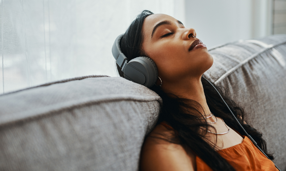 Woman with long dark hair lying back on a sofa while wearing headphones and smiling softly with her eyes closed.